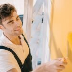 handsome-young-painter-smiling-while-painting-wall-CVNVD59.jpg